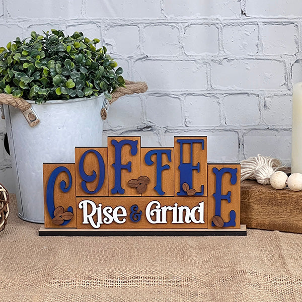 A Coffee Mini Word Block from Janet's Craft Corner, featuring the phrase "Coffee: Rise & Grind" in blue letters with coffee bean decorations, sits on a table. Nearby, a rustic white bucket filled with greenery and a beige candle enhances the home decor against a pristine white brick wall.