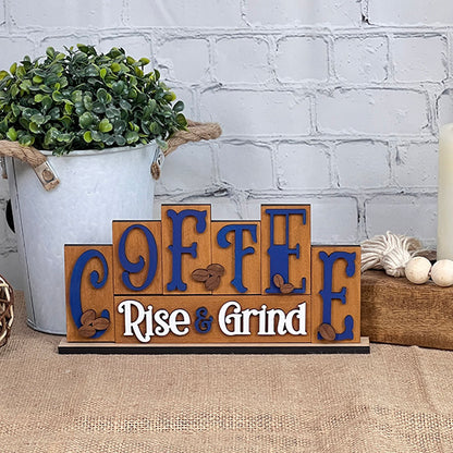 A Coffee Mini Word Block from Janet's Craft Corner, featuring the phrase "Coffee: Rise & Grind" in blue letters with coffee bean decorations, sits on a table. Nearby, a rustic white bucket filled with greenery and a beige candle enhances the home decor against a pristine white brick wall.