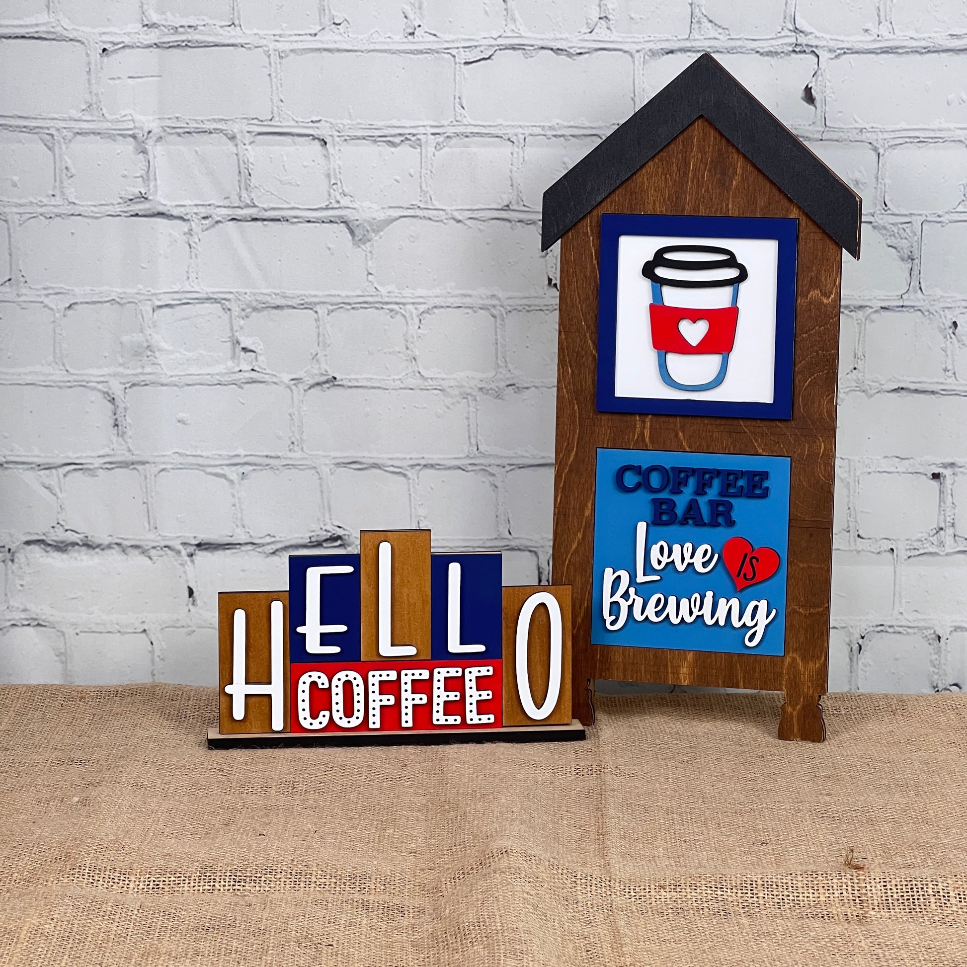 Set against a burlap backdrop and brick wall, the Decorative Coffee Signs by Janet's Craft Corner feature charming designs. The Hello Coffee Mini Word Block displays "HELLO COFFEE" in bold letters, while another sign, shaped like a quaint house, adds charm with phrases "COFFEE BAR" and "Love is Brewing," making it an ideal addition for any DIY kit enthusiast's home decor.