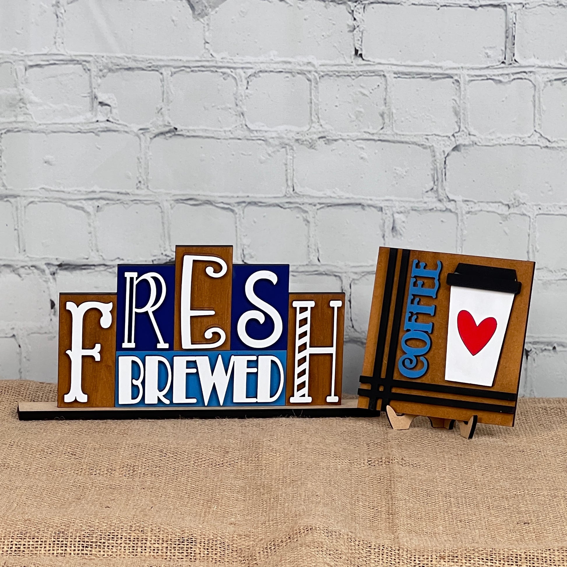 A decorative sign from Janet's Craft Corner, the Fresh Brewed Mini Word Block, features stylized letters spelling out "Fresh Brewed," adding a charming touch to home décor. Next to it is another sign displaying a coffee cup with a red heart and the word "coffee" in blue. These elements are set against a white brick wall backdrop, completing this cozy DIY kit scene.