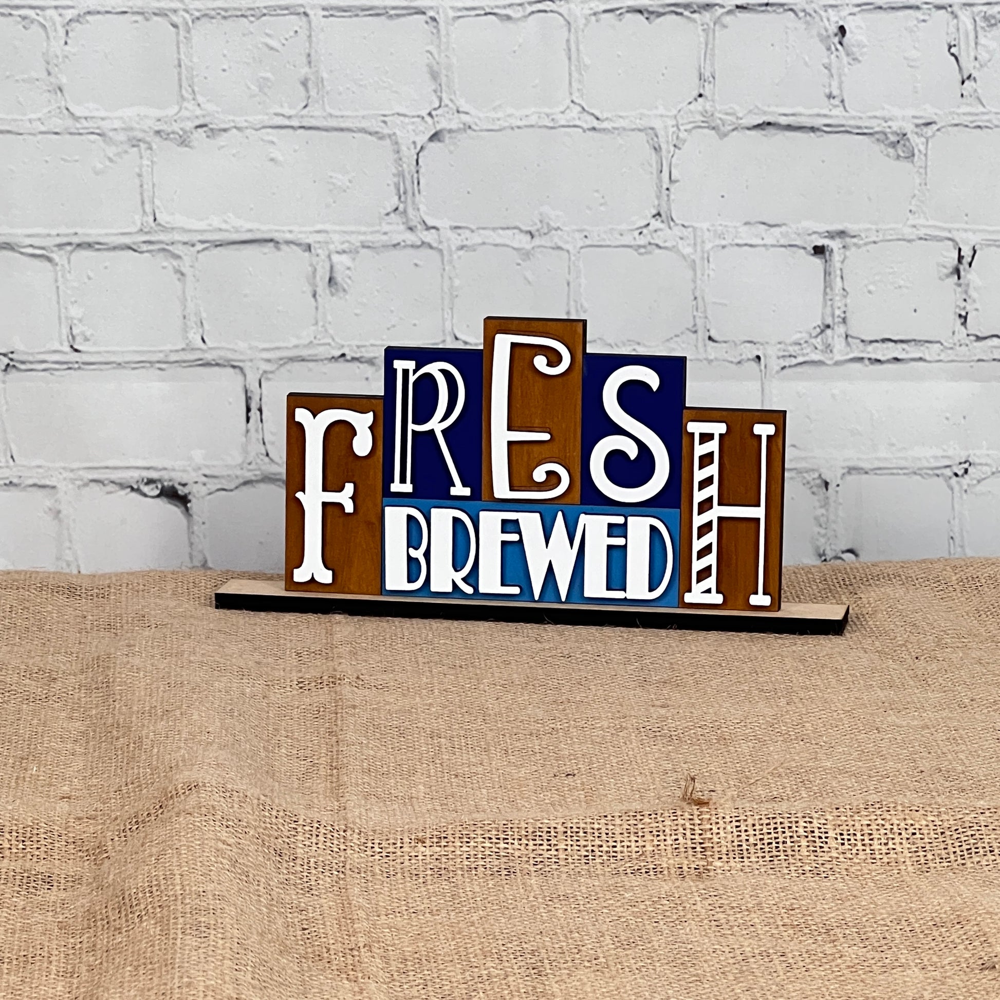 Introducing the Fresh Brewed Mini Word Block from Janet's Craft Corner, a delightful home décor piece. This DIY kit showcases a decorative sign featuring the words "Fresh Brewed" in an assortment of fonts and colors set against a wooden backdrop. It is elegantly displayed on a burlap-covered surface with a chic white brick wall providing the perfect background.