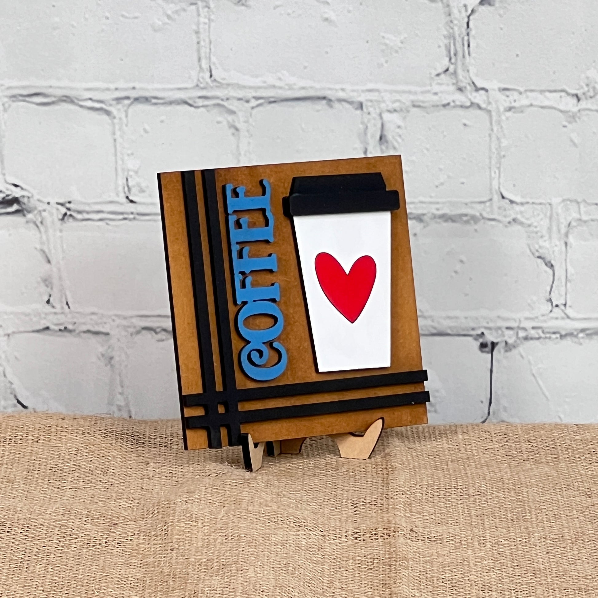 Celebrate coffee lovers with Janet's Craft Corner Coffee Swappable Squares—a DIY seasonal home decor kit featuring a white cup with a red heart, blue "COFFEE" text, and black lid on a brown striped background. Perfect handmade charm!.