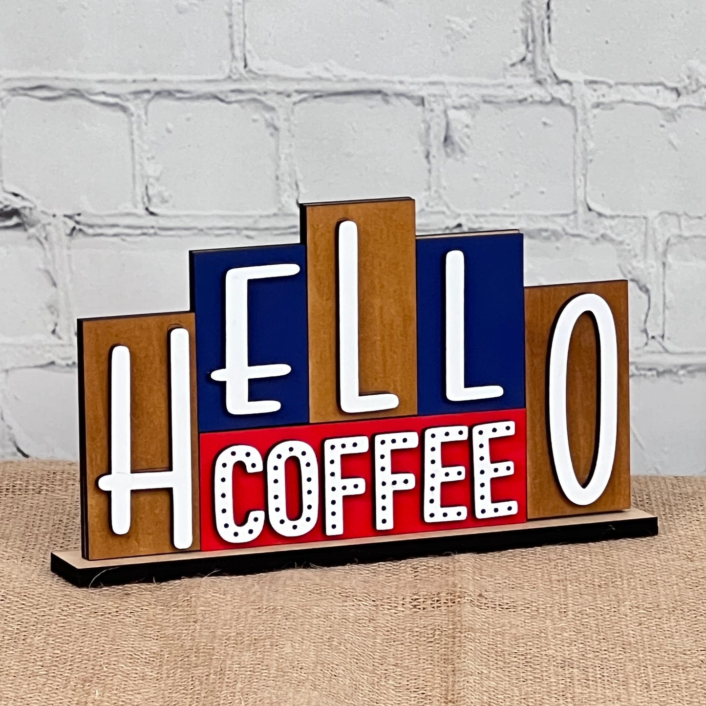 Enhance your home decor with the "Hello Coffee Mini Word Block" from Janet's Craft Corner. Featuring vivid white and red lettering against a striking blue and red background on a burlap-covered surface, this charming piece is perfect for your next DIY kit project set against a white brick wall.