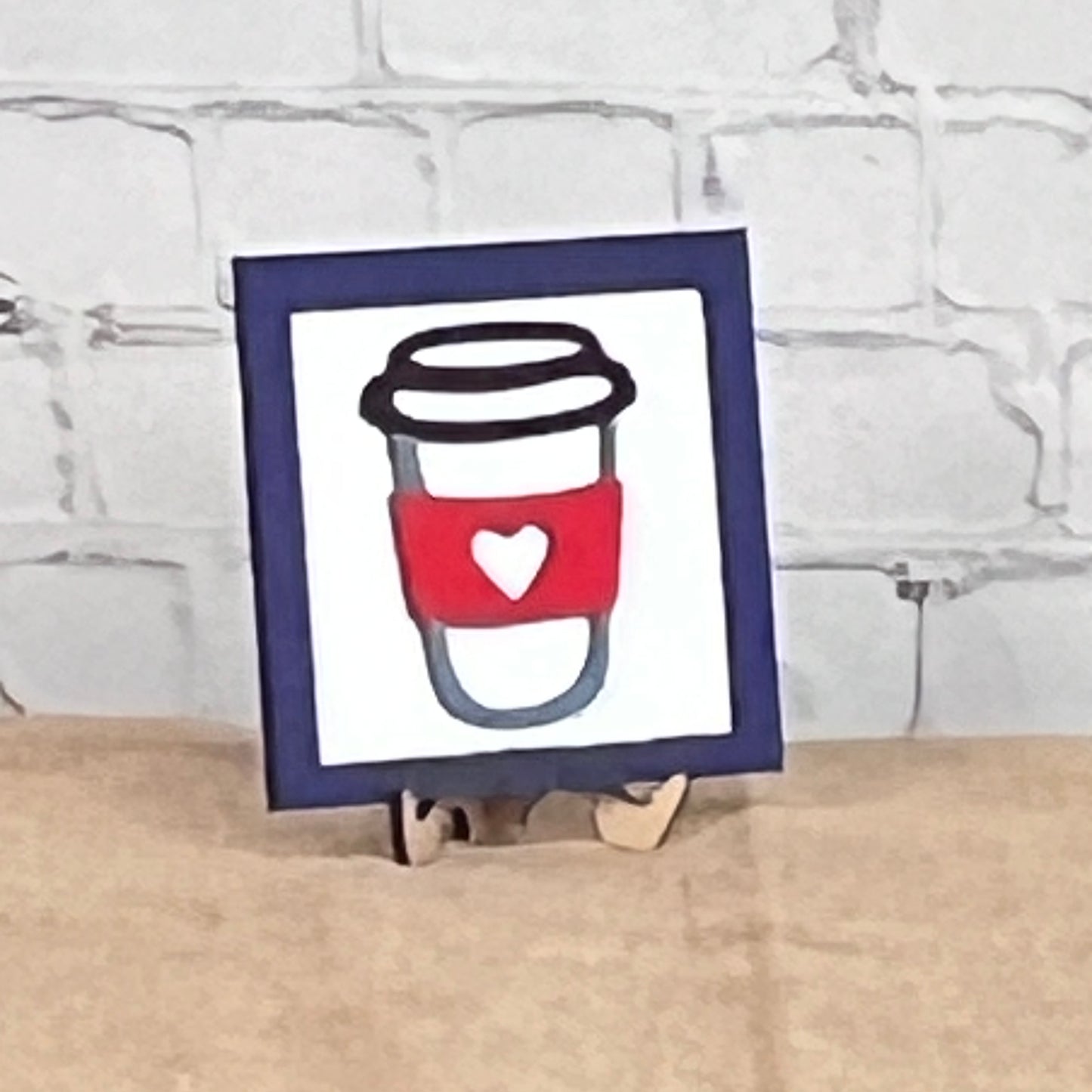 Coffee Squares by Janet's Craft Corner: a framed drawing of a coffee cup with a red sleeve and white heart, displayed on wood risers as charming DIY home decor against a white brick wall.