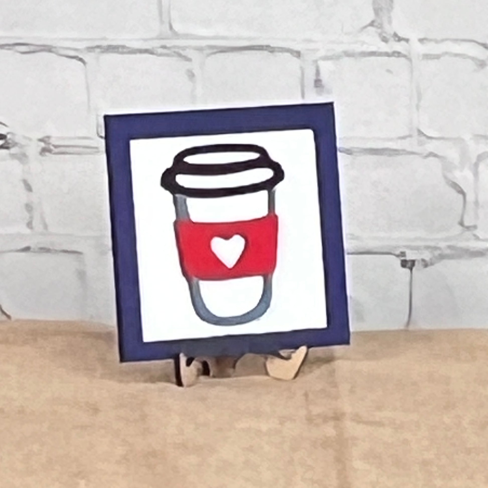 Coffee Squares by Janet's Craft Corner: a framed drawing of a coffee cup with a red sleeve and white heart, displayed on wood risers as charming DIY home decor against a white brick wall.