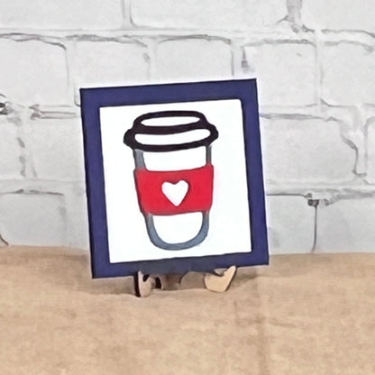 Coffee Squares by Janet's Craft Corner: a framed drawing of a coffee cup with a red sleeve and white heart, displayed on wood risers as charming DIY home decor against a white brick wall.