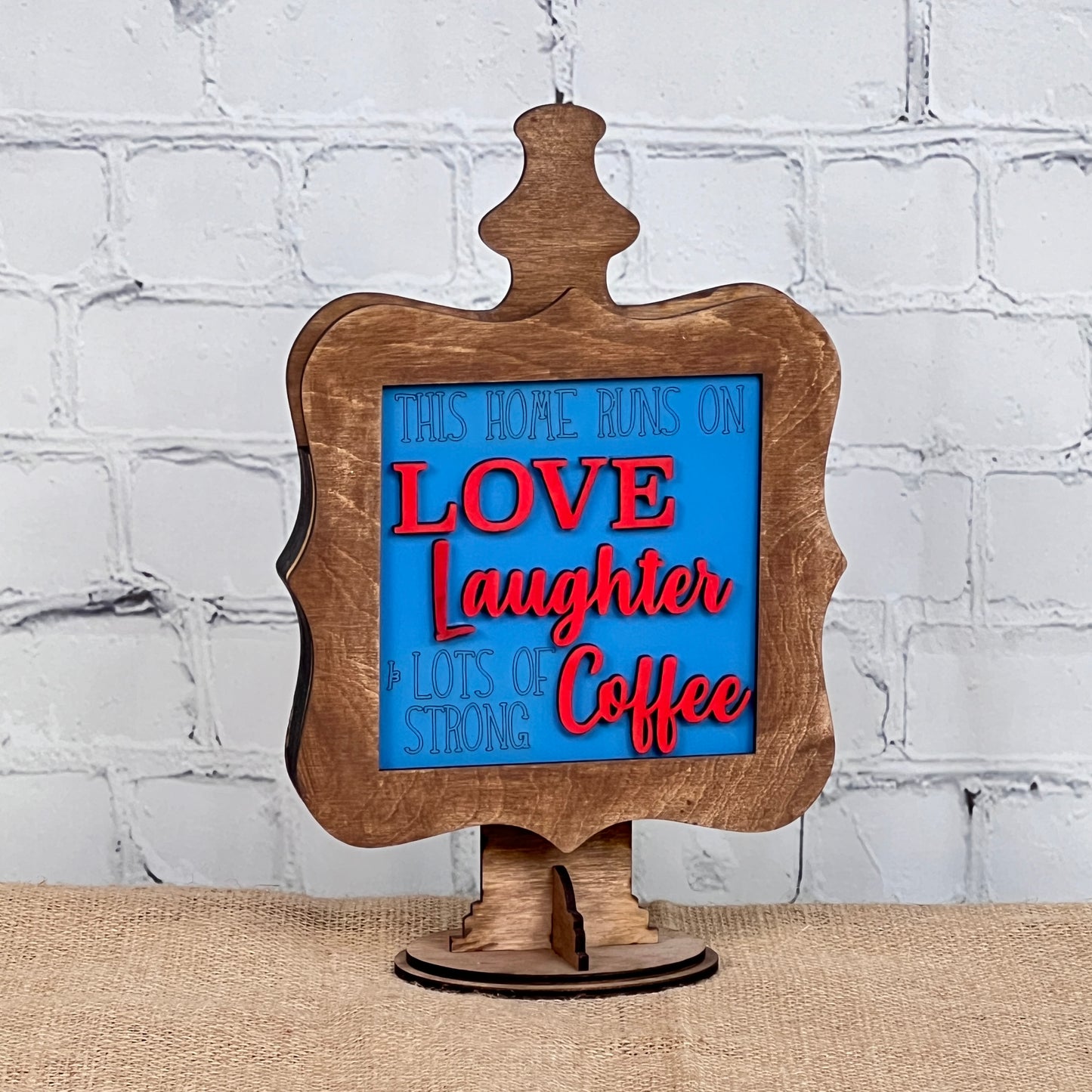 Janet's Craft Corner presents Coffee Squares—a decorative wooden sign with a blue and red message, "This home runs on love, laughter & lots of strong coffee," ideal for DIY home decor on burlap or white brick backgrounds.
