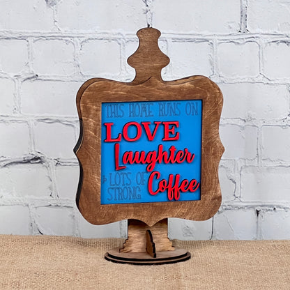 Janet's Craft Corner presents Coffee Squares—a decorative wooden sign with a blue and red message, "This home runs on love, laughter & lots of strong coffee," ideal for DIY home decor on burlap or white brick backgrounds.