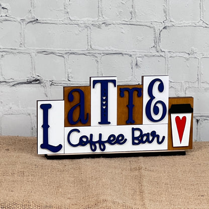 This charming home décor piece from Janet's Craft Corner, called the Latte Mini Word Block, displays "Latte Coffee Bar" in blue and black on a white background. It includes a delightful coffee cup illustration with a red heart, making it perfect for coffee lovers and adding warmth to any space when displayed against its white brick wall setting.