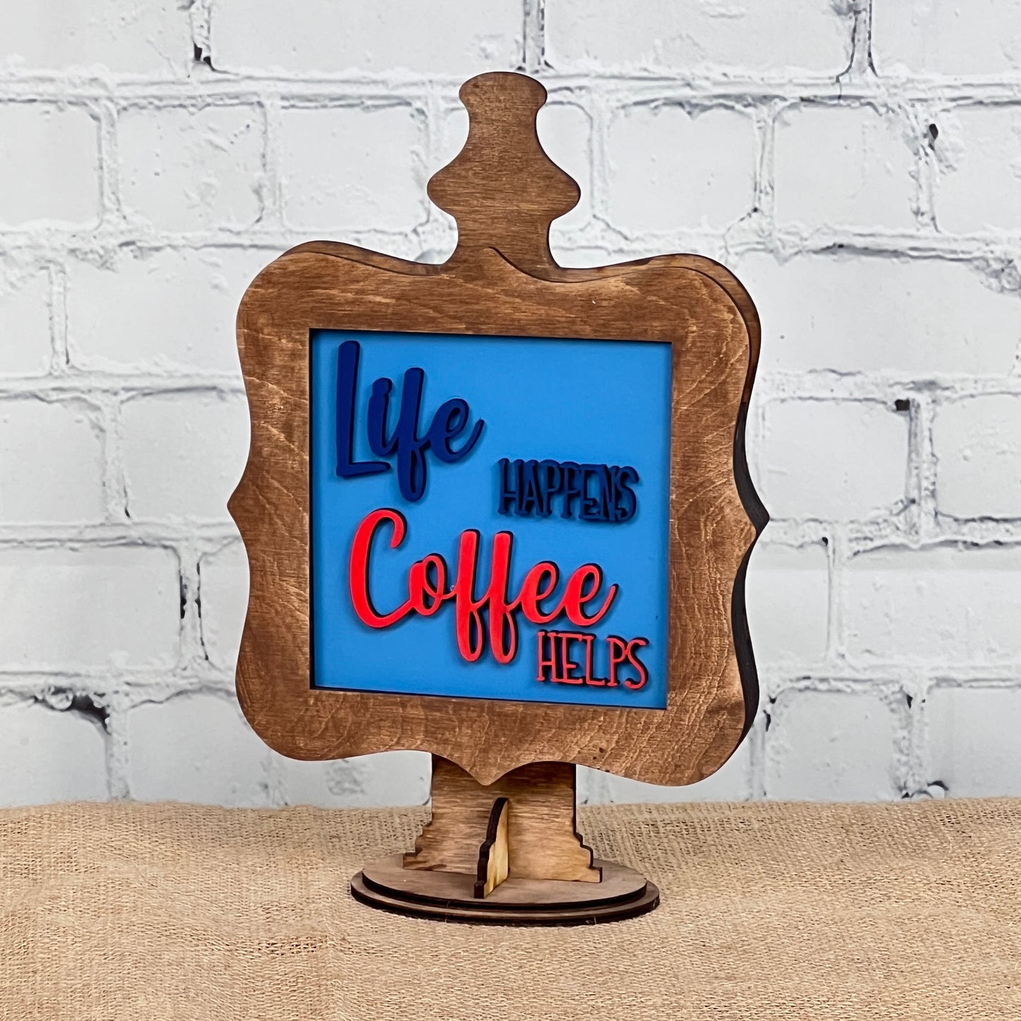 The Coffee Squares tabletop sign from Janet's Craft Corner features blue and red raised letters on a blue background reading "Life happens, coffee helps," making it an ideal DIY home decor accent for any space.