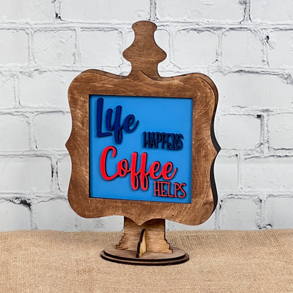 The Coffee Squares tabletop sign from Janet's Craft Corner features blue and red raised letters on a blue background reading "Life happens, coffee helps," making it an ideal DIY home decor accent for any space.
