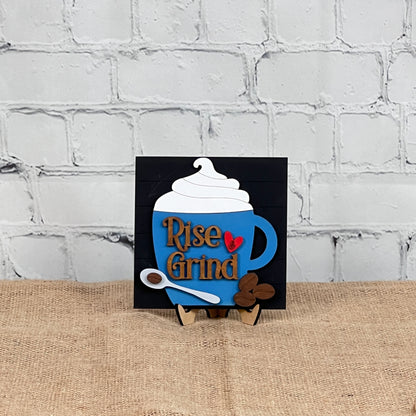 The Coffee Swappable Squares DIY seasonal home decor craft kit by Janet's Craft Corner features a blue coffee mug sign with whipped cream, "Rise & Grind" text, and a red heart—perfect decor for coffee lovers.