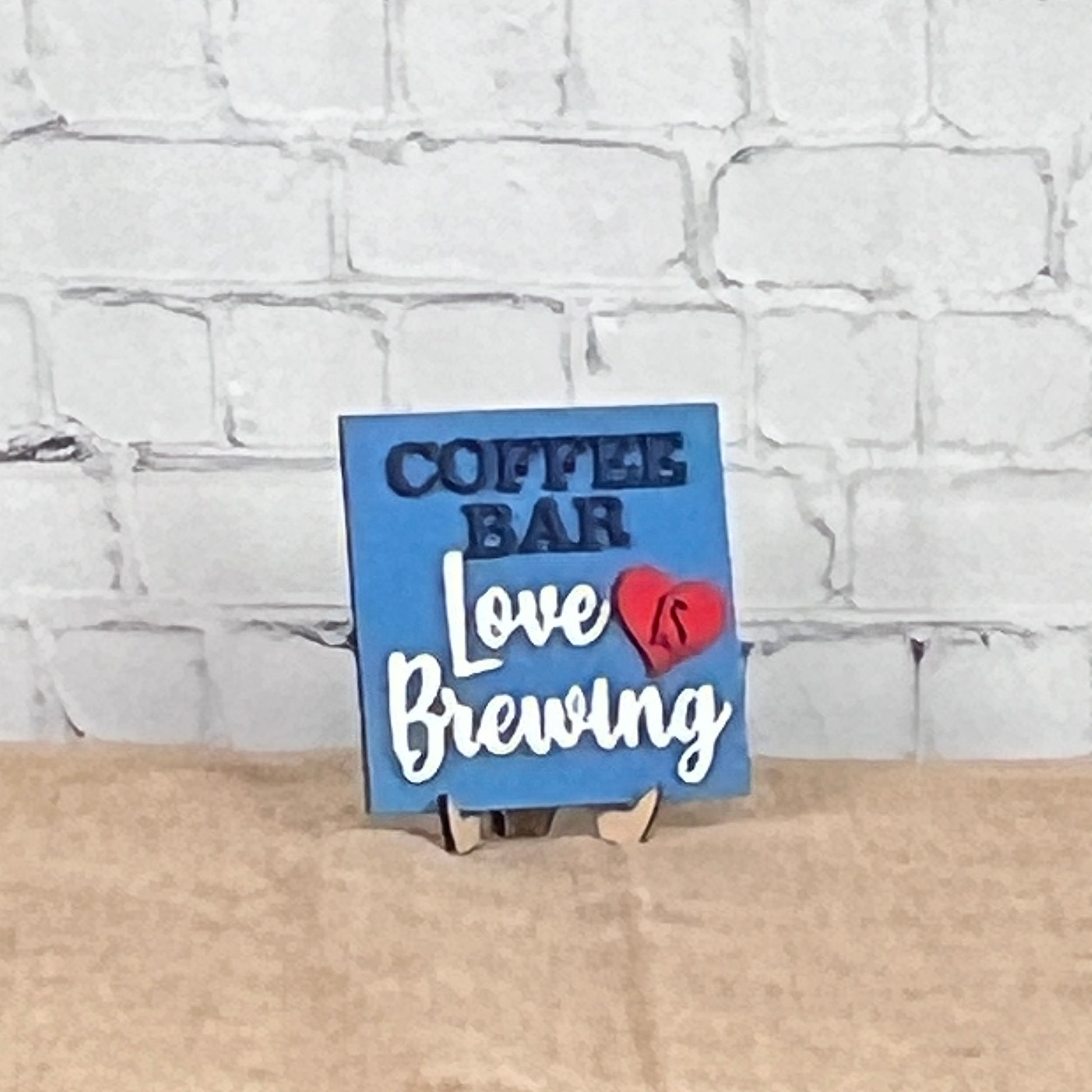 A small blue sign from Janet’s Craft Corner’s Coffee Squares collection sits on a tan surface against a white brick wall, reading "COFFEE BAR Love IS Brewing" with "IS" inside a red heart—perfect for cozy DIY home decor.