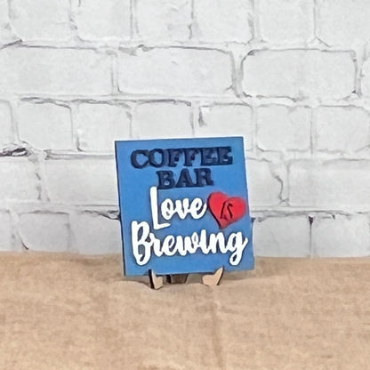 A small blue sign from Janet’s Craft Corner’s Coffee Squares collection sits on a tan surface against a white brick wall, reading "COFFEE BAR Love IS Brewing" with "IS" inside a red heart—perfect for cozy DIY home decor.