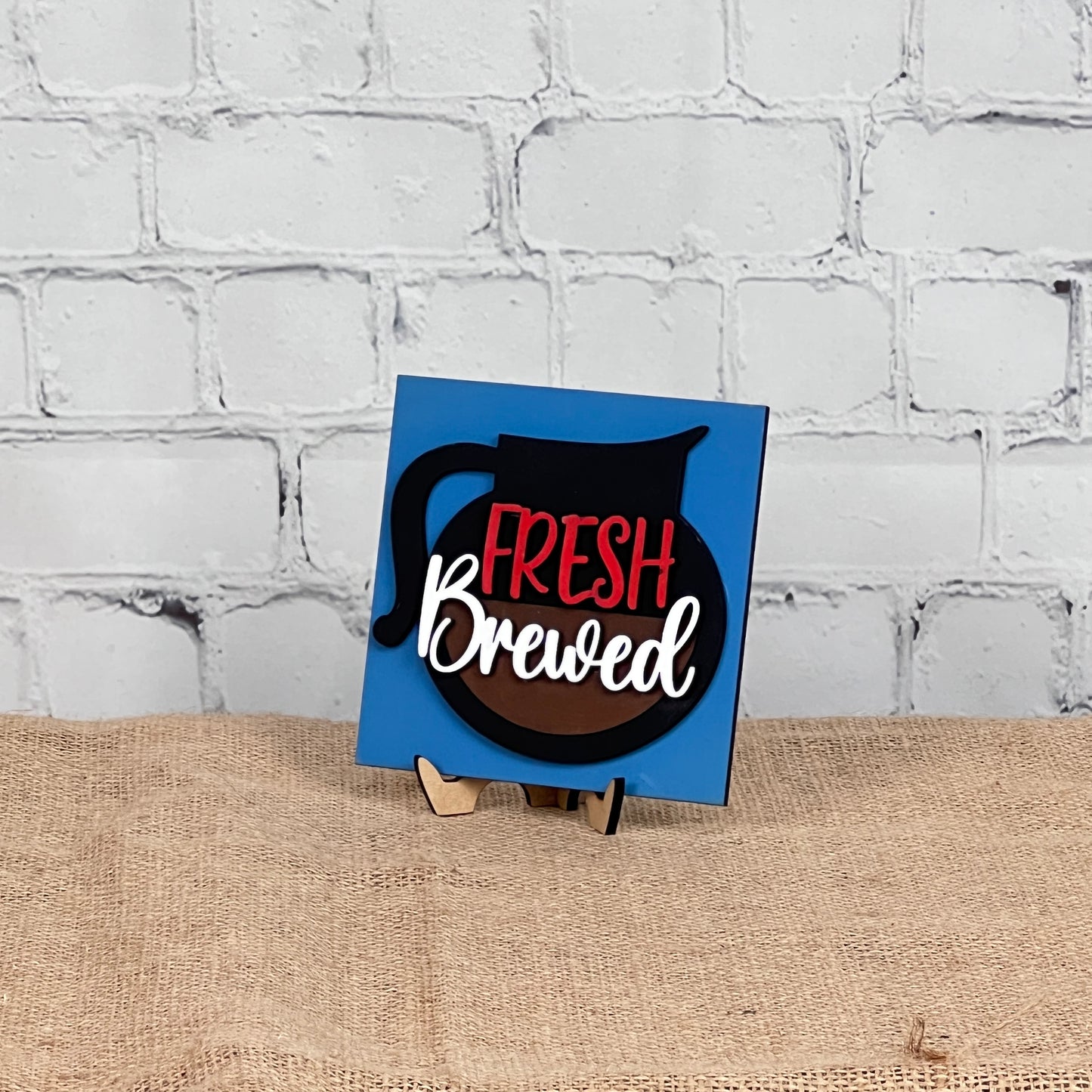 A blue square sign from Janet's Craft Corner's Coffee Swappable Squares DIY seasonal home decor kit displays a coffee pot graphic and "Fresh Brewed" in red and white, on a wooden stand against a white brick wall and burlap surface.