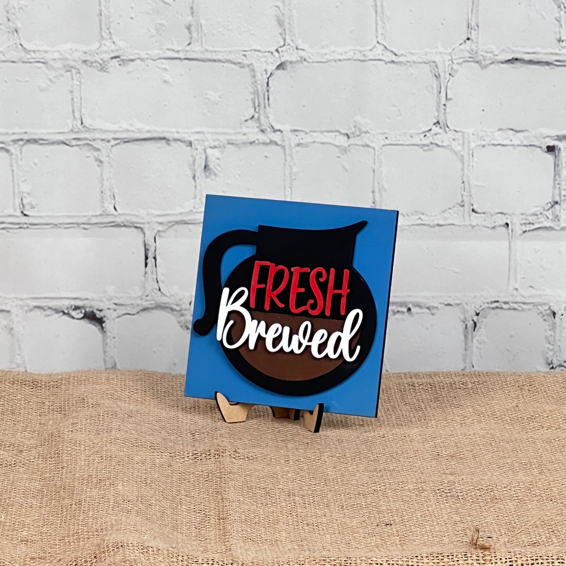 A blue square sign from Janet's Craft Corner's Coffee Swappable Squares DIY seasonal home decor kit displays a coffee pot graphic and "Fresh Brewed" in red and white, on a wooden stand against a white brick wall and burlap surface.