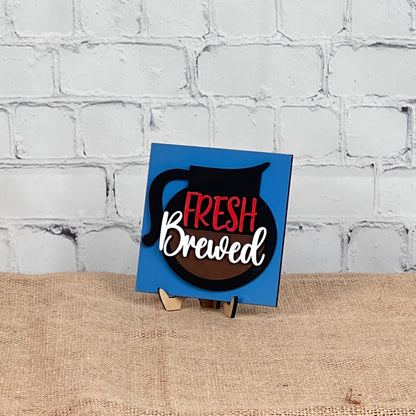 A blue square sign from Janet's Craft Corner's Coffee Swappable Squares DIY seasonal home decor kit displays a coffee pot graphic and "Fresh Brewed" in red and white, on a wooden stand against a white brick wall and burlap surface.