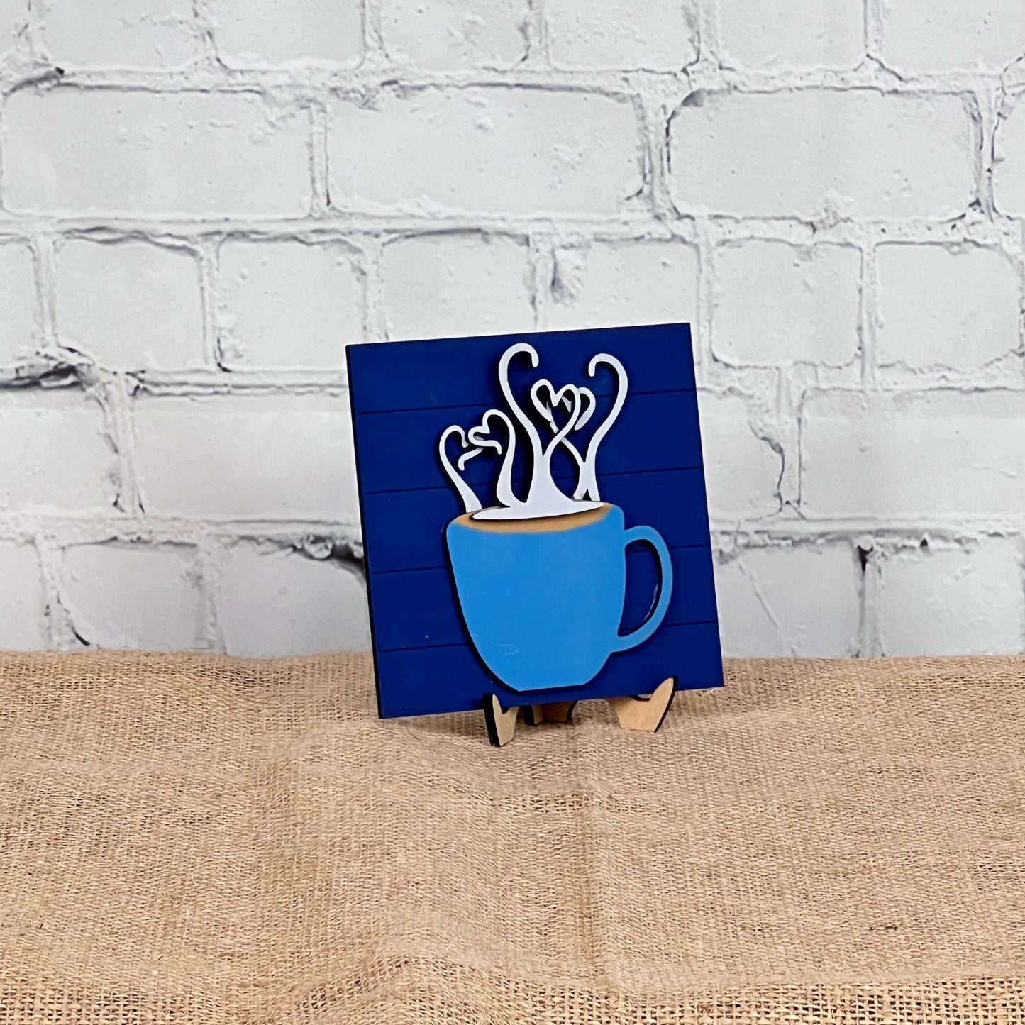 A blue mug with heart-shaped steam appears on Janet's Craft Corner's Coffee Swappable Squares— a DIY seasonal home decor kit—displayed on a small easel over burlap, set against a white brick wall for cozy charm.