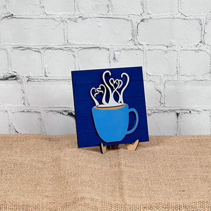 A blue mug with heart-shaped steam appears on Janet's Craft Corner's Coffee Swappable Squares— a DIY seasonal home decor kit—displayed on a small easel over burlap, set against a white brick wall for cozy charm.