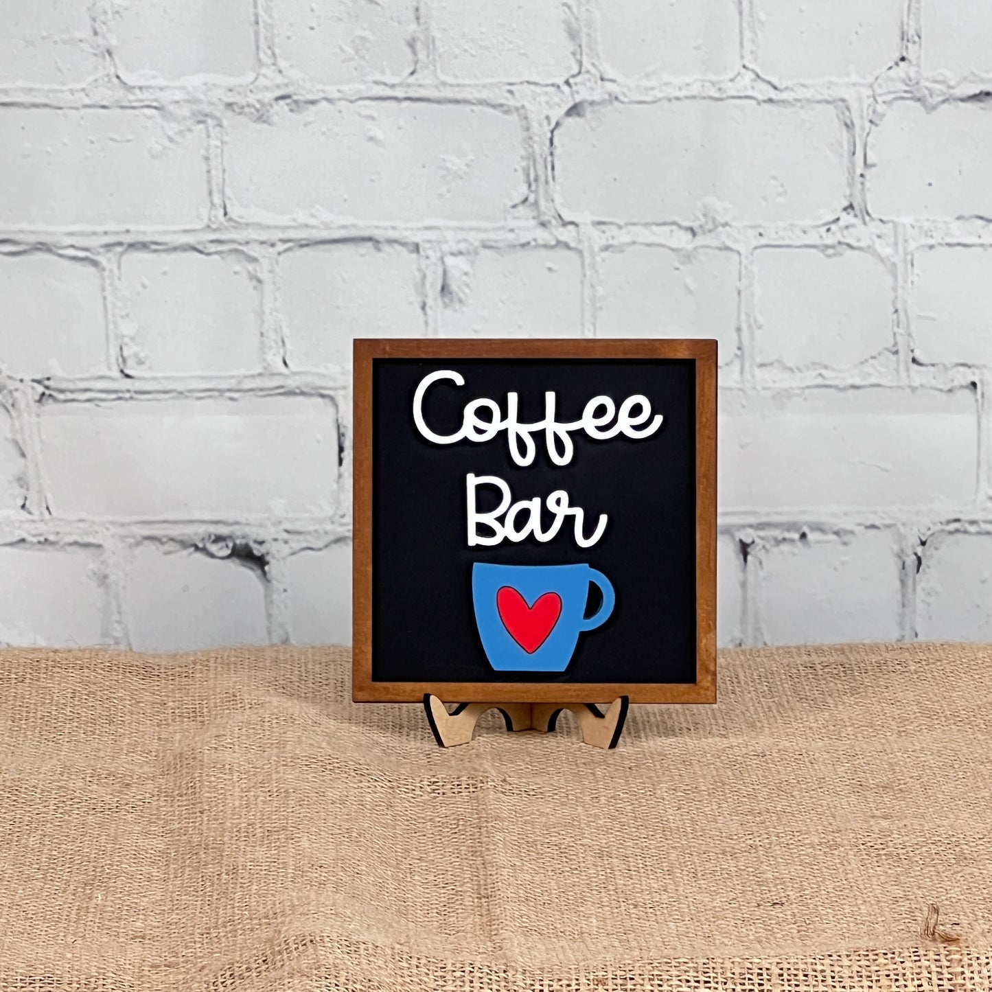 A small sign reading "Coffee Bar" with a blue coffee cup and red heart sits on a stand—perfect inspiration for your next Coffee Swappable Squares DIY kit from Janet’s Craft Corner. Display in front of brick or burlap for cozy charm!.