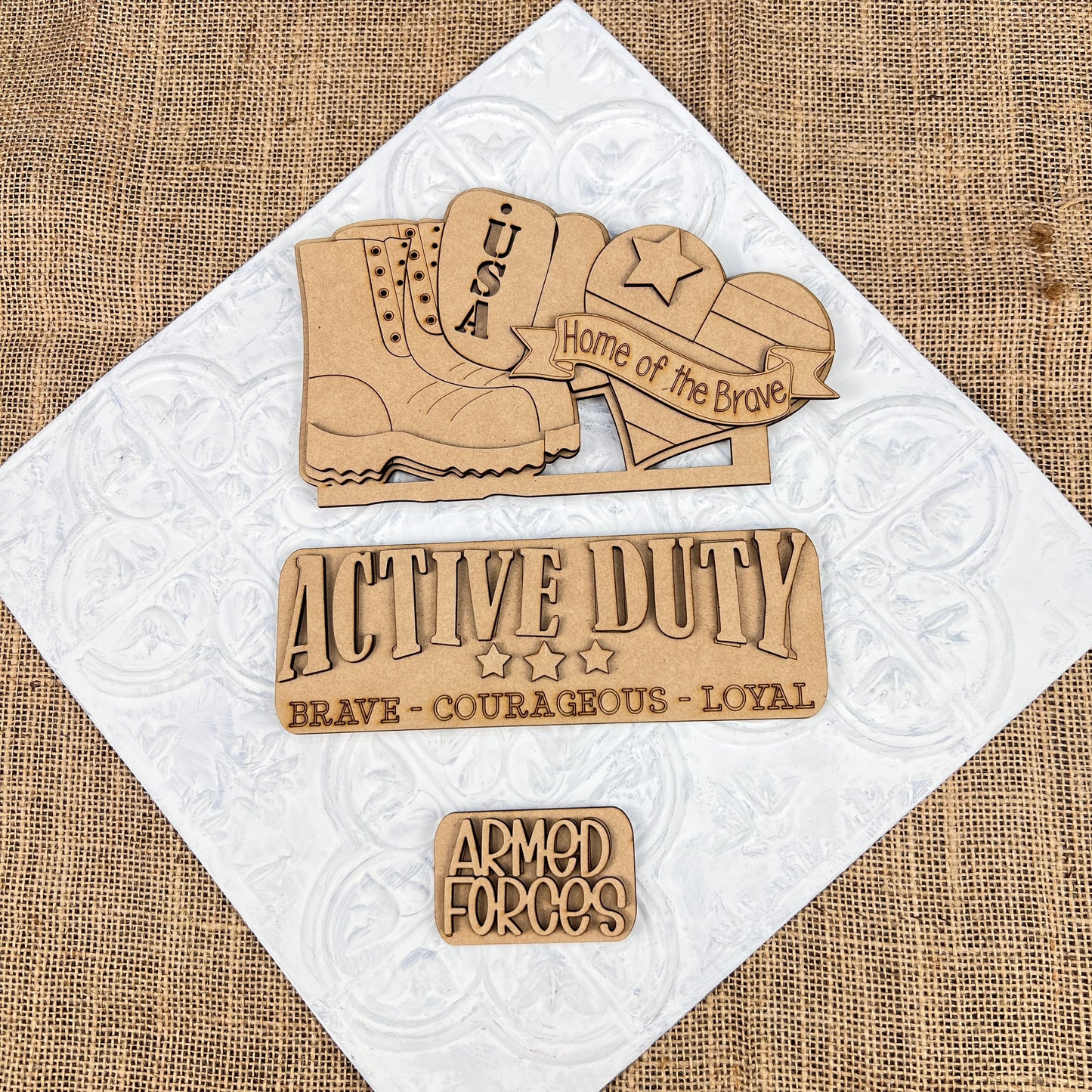 Janet's Craft Corner presents the Military Interchangeable Insert, a DIY home decor kit that showcases wooden cutouts with military themes on a textured background. The set includes items such as a boot, dog tags, and cap emblazoned with "USA" and "Home of the Brave." It features phrases like "Active Duty" and "Armed Forces," making it perfect for creating interchangeable patriotic displays in your home.