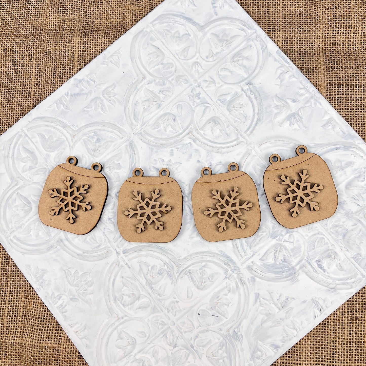 Five charming ornaments from the Cup of Cheer Hot Cocoa Decor Set by Janet's Craft Corner are beautifully showcased on a textured white tile surface with a burlap background, adding delightful charm to your home decor.