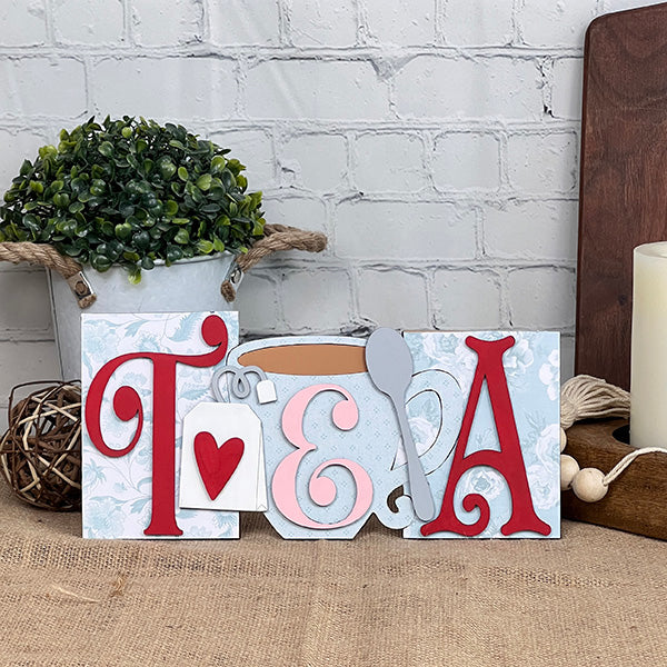 Enhance your home décor with the delightful Tea Word Block from Janet's Craft Corner, a wooden decorative sign displaying the word "TEA" in striking red and white hues. This charming piece features a heart, teacup, and spoon motifs and is beautifully set against a white brick wall alongside a small plant, wicker ball, and candles. Perfect for adding a touch of charm to any space!