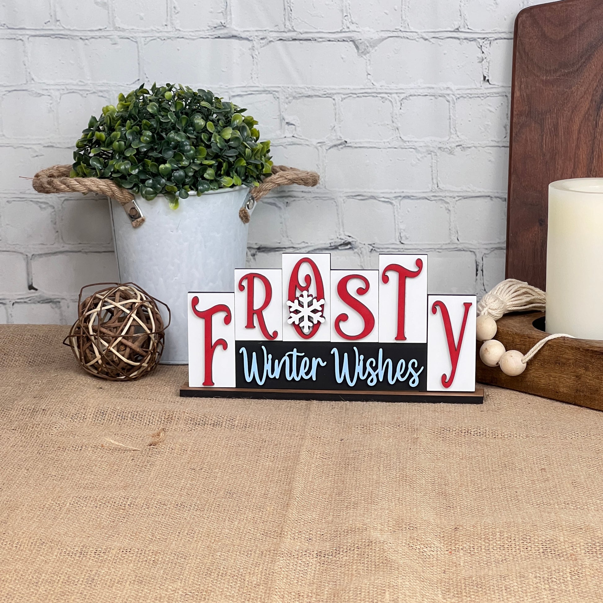 The Frosty Mini Word Block Shelf Sitter from Janet's Craft Corner is a delightful DIY decor kit that features "Frosty Winter Wishes" in red and white letters with a charming snowflake design. It brings a festive flair to your home decor when placed on a burlap-covered surface next to a white pot filled with greenery, accompanied by a woven ball and candle on a wooden tray against the backdrop of a brick wall.