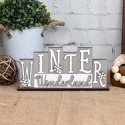 The "Winter Wonderland Mini Word Block Shelf Sitter" from Janet's Craft Corner adorns a white brick wall, featuring snowflake accents. It rests on burlap alongside a potted plant, woven sphere, and wooden beads—creating the perfect home décor piece that adds warmth and charm.