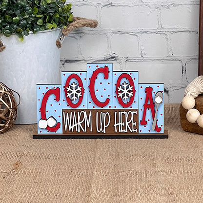 The Cocoa Mini Word Block Shelf Sitter from Janet's Craft Corner features the phrase "COCOA WARM UP HERE" in bold red letters on a light blue background adorned with snowflakes and marshmallows. This DIY Decor Kit is ideal for adding a personalized touch to your home. It can be displayed on burlap beside a potted plant and woven ball, set against a white brick wall for an inviting look.