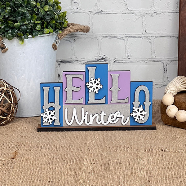 Introducing the "Hello Winter" Mini Word Block Shelf Sitter by Janet's Craft Corner. This DIY decor kit features blue, purple, and gray blocks with snowflake-adorned letters. It is showcased on a burlap-covered surface, complete with greenery and wooden beads, making it an ideal addition to your home décor collection.