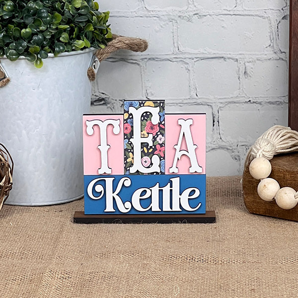 The Tea Kettle Mini Word Block from Janet's Craft Corner features the word "TEA" prominently against a floral background, with "Kettle" below. This delightful piece, ideal for home décor enthusiasts or as a DIY kit project, is displayed on a burlap surface surrounded by plants and rustic elements.