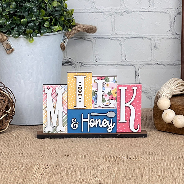 The Janet's Craft Corner Milk & Honey Mini Word Block features vibrant patterned blocks set against a burlap backdrop. It's an ideal choice for home décor enthusiasts, complete with a potted plant and decorative balls in the background. Perfect as a DIY kit, it brings charm and creativity to any space.