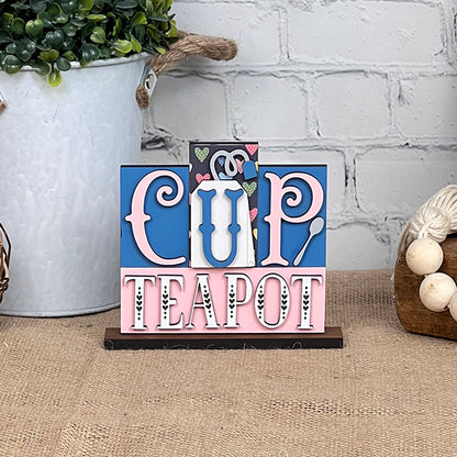 Perfect for tea enthusiasts, Janet's Craft Corner offers the Cup-Teapot Mini Word Block DIY Kit. It features the words "Cup" and "Teapot" in bright letters, accompanied by hearts and a teapot design. The set is displayed on textured brick wall material with burlap, creating a cozy aesthetic alongside plant and yarn decorations.