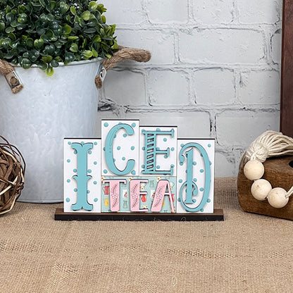 The delightful Iced Tea Mini Word Block from Janet's Craft Corner brings a burst of color with its whimsical letters on burlap. When placed among rustic decorations and a potted plant, it beautifully complements your home décor against the white brick background, making it ideal for DIY kit enthusiasts!