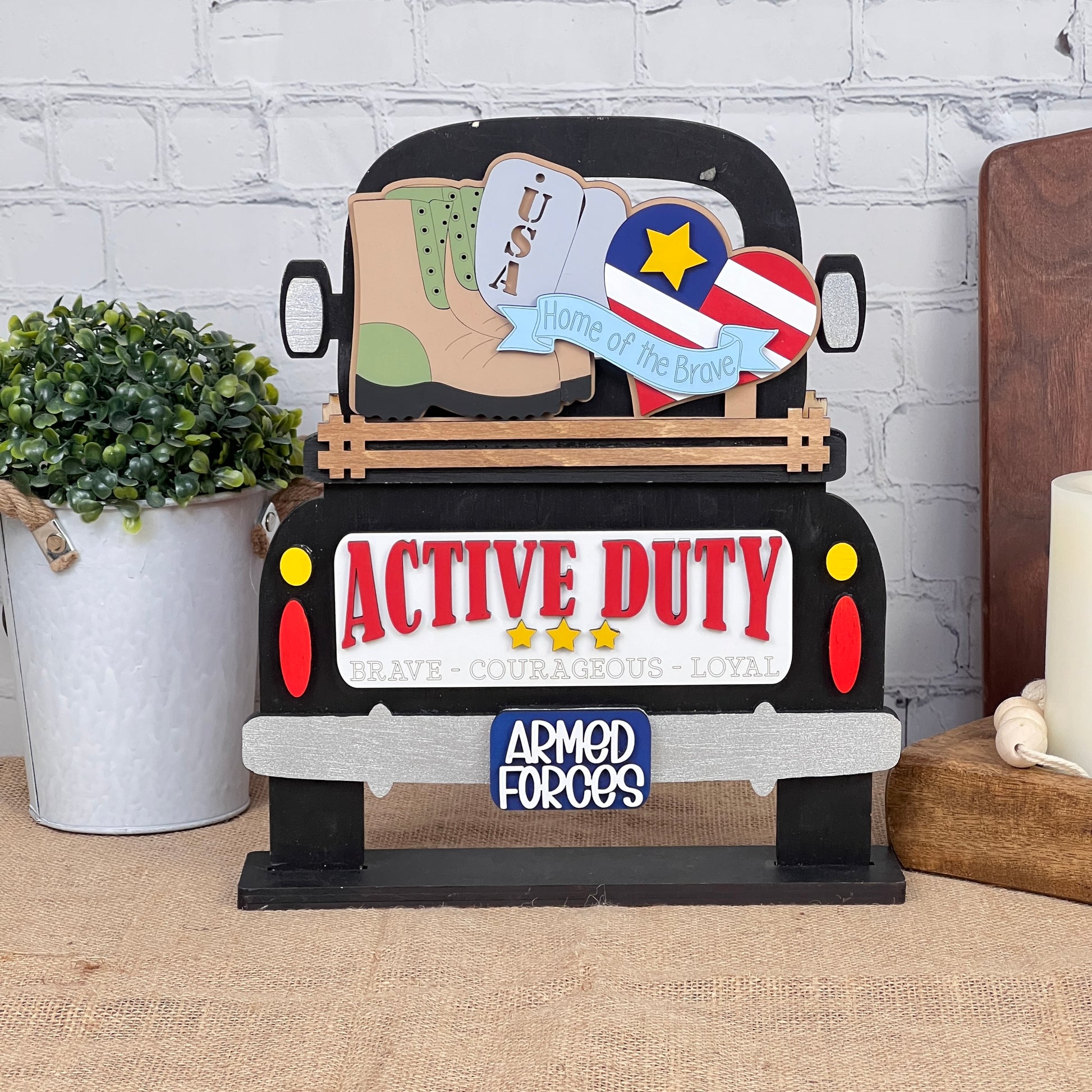 A charming truck-shaped sign labeled "Active Duty," adorned with military service symbols like boots, a star, and a heart inscribed with "Home of the Brave," is part of Janet's Craft Corner's Military Interchangeable Insert DIY home decor kit. The display also includes a small plant and candle on the table.