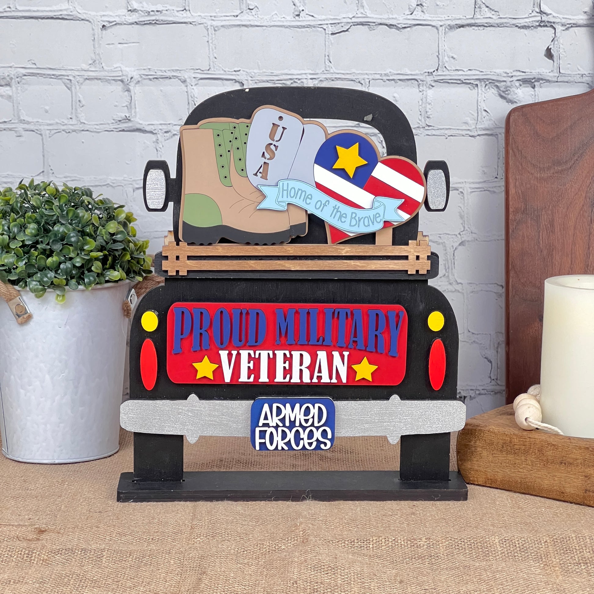 Introducing the Military Interchangeable Insert from Janet's Craft Corner, a DIY home decor kit that features decorative wooden artwork with a truck adorned with "Proud Military Veteran" and "Armed Forces" signs. This piece includes patriotic symbols such as a heart, star, and boots labeled with "USA," along with the inspiring text "Home of the Brave." It's an ideal choice for interchangeable home decor, beautifully displayed against a brick wall alongside a plant and candle.