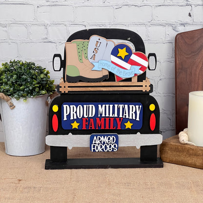 The Military Interchangeable Insert by Janet's Craft Corner is a decorative sign that showcases a vintage truck adorned with the phrases "Proud Military Family" and "Armed Forces." The truck bed features a boot, USA sign, and heart with flag design. This DIY home decor kit comes equipped with elements like a potted plant, candle, and brick wall to enhance your patriotic display at home.