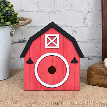 The Barn Interchangeable Set by Janet's Craft Corner is a decorative red birdhouse styled like a barn. It features a round white entrance and black roof accents, set against a brick wall backdrop. This charming piece of home décor is beautifully complemented by a potted plant and wooden block, infusing any space with rustic charm.