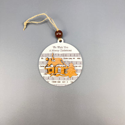 Janet's Craft Corner presents an exquisite Christmas ornament called Sheet Music Ornaments, featuring a rustic circular wooden design with a charming house and trees. The background is beautifully embellished with sheet music detailing the phrase "We Wish You A Merry Christmas," making it an ideal gift for music lovers. It hangs gracefully thanks to a string and bead.