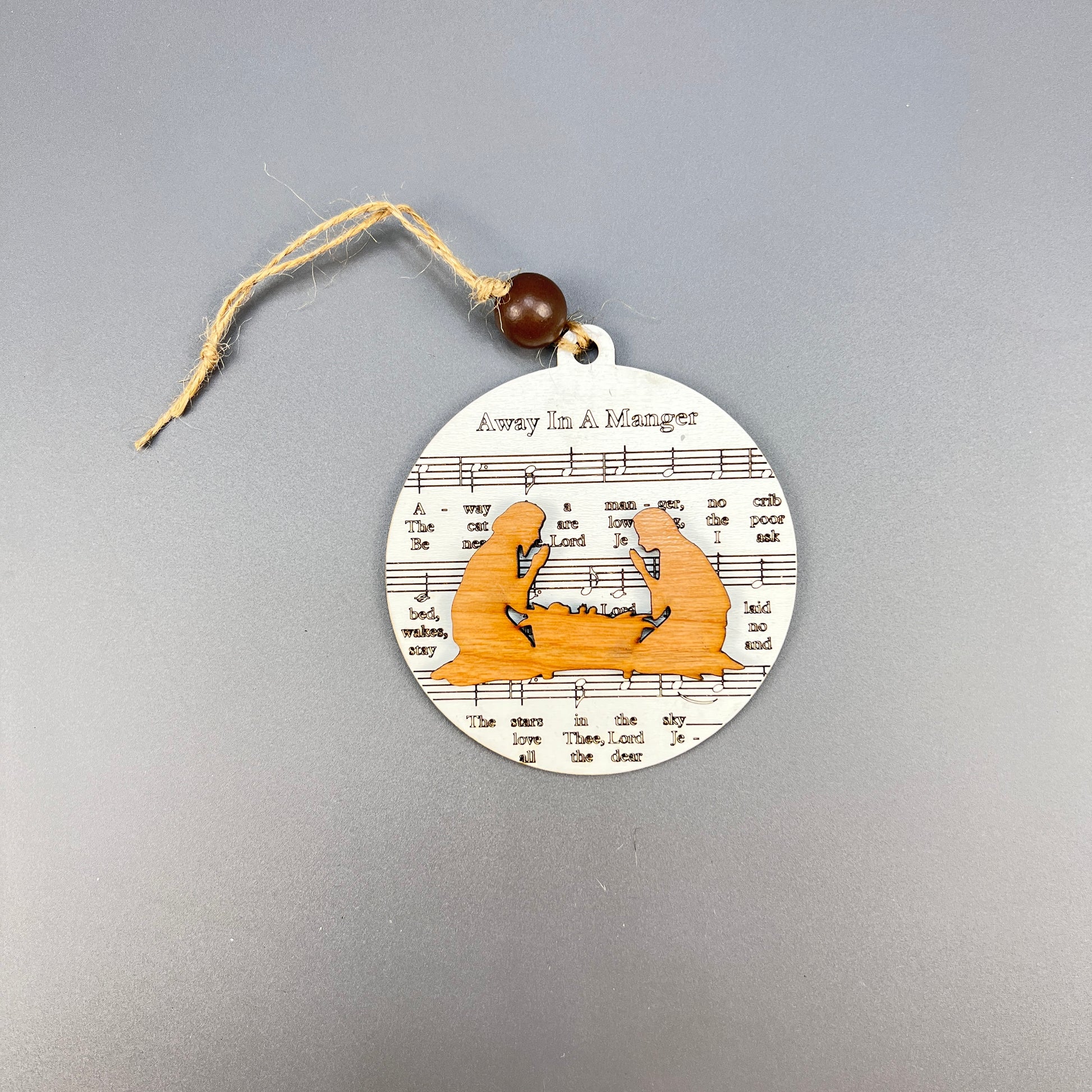 The Sheet Music Ornaments by Janet's Craft Corner exude rustic charm, featuring a round ornament with a beautifully cut-out nativity scene over sheet music for "Away in a Manger." Ideal for music lovers, it includes a string and bead for effortless hanging.