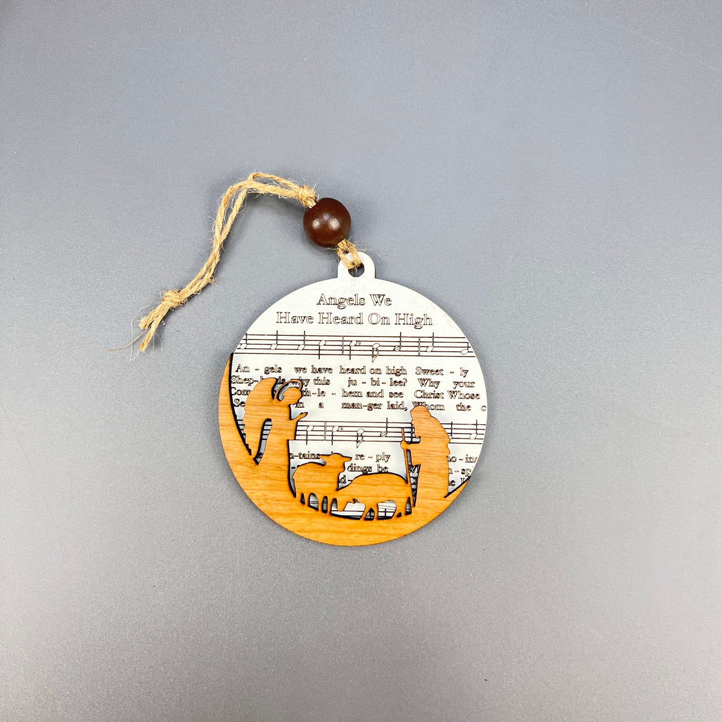 Introducing Janet's Craft Corner Sheet Music Ornaments: This ornament embodies rustic charm with a silhouette of the nativity scene featuring Mary, Joseph, and baby Jesus. The background showcases sheet music for "Angels We Have Heard On High," making it an ideal choice for music enthusiasts. It includes a twine and bead loop for easy hanging.