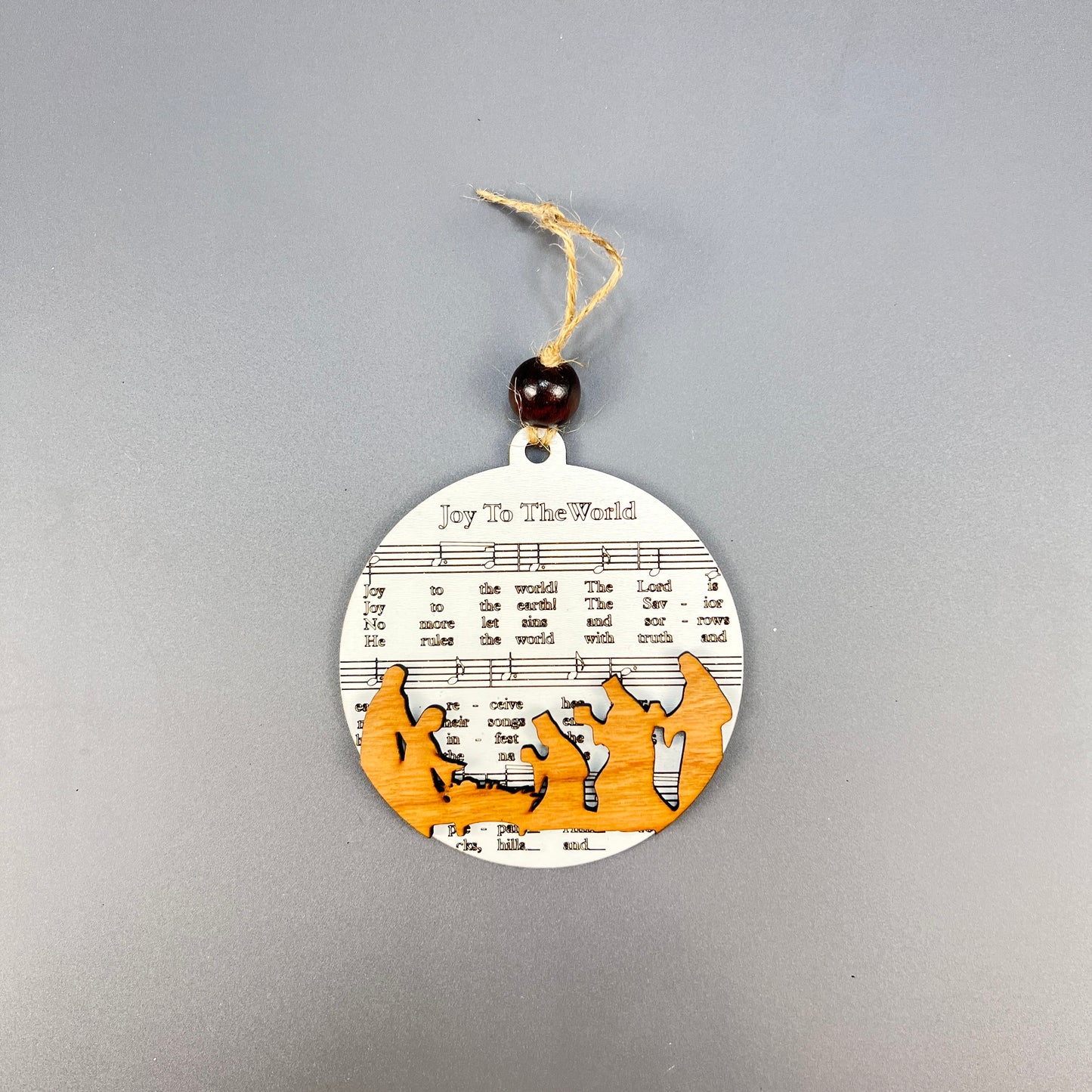 Introducing Janet's Craft Corner's Sheet Music Ornaments – a rustic round Christmas ornament that beautifully captures silhouettes of the Nativity scene, set against a background of musical notes and lyrics from "Joy to the World." Ideal for music lovers, it comes with a string and bead for effortless hanging.