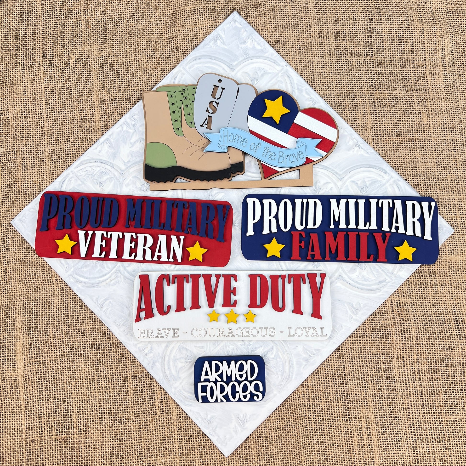 Showcasing military-themed crafts on a burlap background, this DIY home decor kit includes an interchangeable display that features a USA sign with a boot and the text "Home of the Brave." Created by Janet's Craft Corner, this hand-painted Military Interchangeable Insert also has badges reading "Proud Military Veteran" and "Proud Military Family.