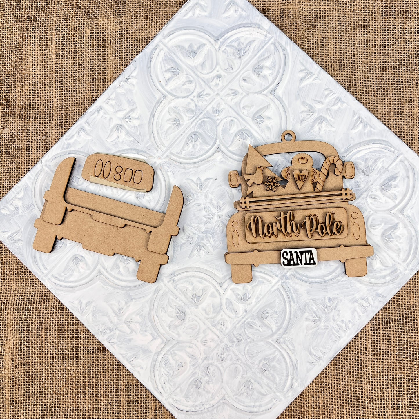 Displayed on a textured white and burlap background are two wooden cutouts of vintage trucks. One is an Antique Truck Ornament from Janet's Craft Corner, featuring "00 800" on the back, while the other displays "North Pole," adorned with Christmas items and a "Santa" license plate. These are perfect additions to your DIY decoration kits or Christmas décor collection.