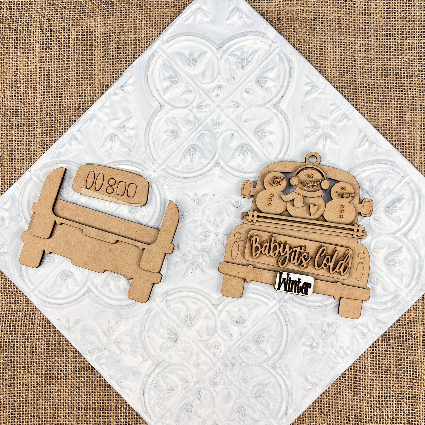 Two antique truck ornaments from Janet's Craft Corner rest on a decorative white tile. One showcases "Baby's Cold Winter" with snowmen, while the other offers a customizable blank section and features an engraving of "0800." These DIY decoration kits are perfect for Christmas décor, adding charm to any burlap background.