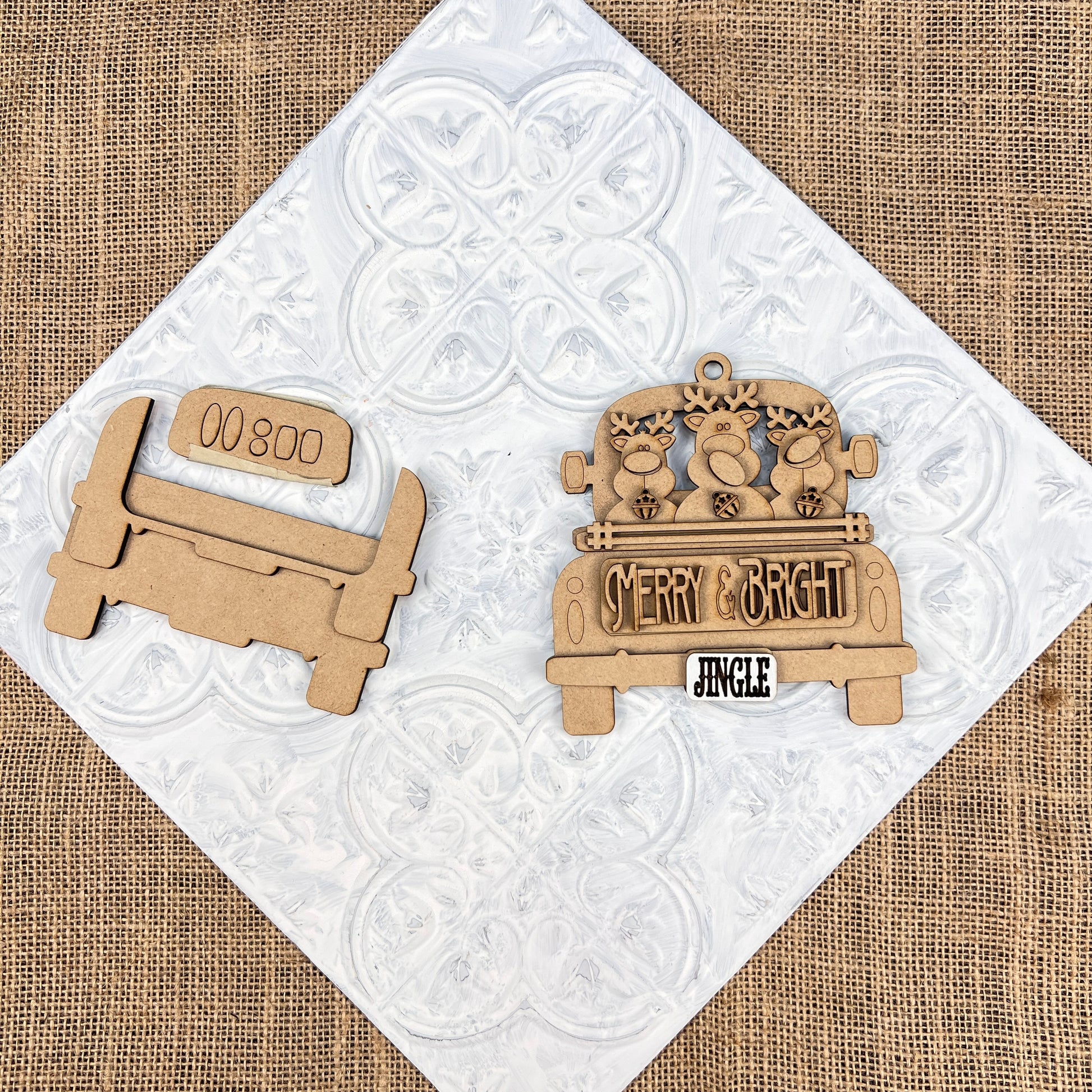 Two wooden cutouts from Janet's Craft Corner add charm to a burlap and white patterned background. One features an antique truck ornament design with the words "Merry & Bright" and "Jingle," accompanied by three reindeer heads, while the other resembles a bench marked with "00800." These are perfect for your DIY Christmas décor projects.