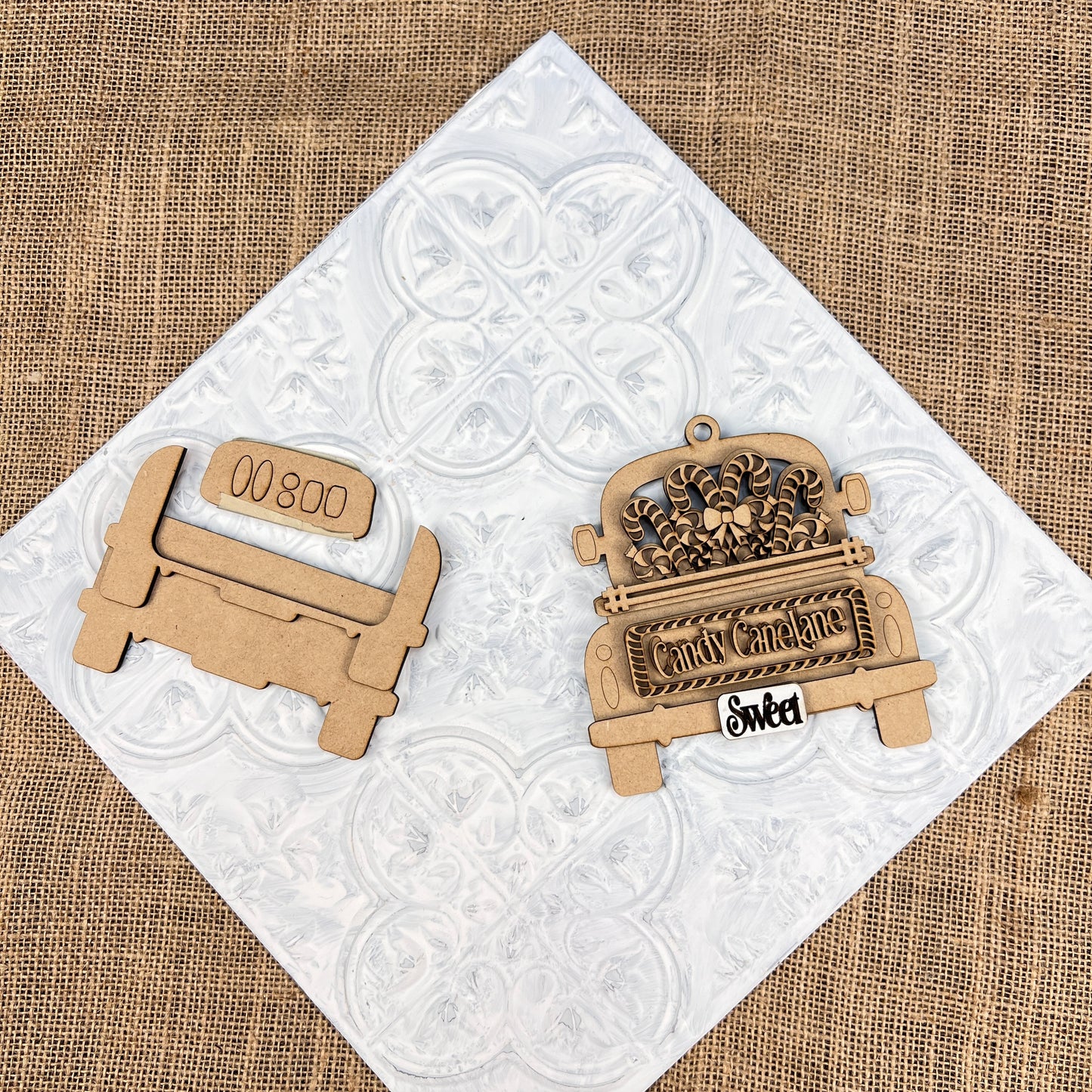 Wooden Christmas ornaments rest elegantly on a textured white and burlap background. The Antique Truck Ornament from Janet's Craft Corner features the delightful phrases "Candy Cane Lane" and "Sweet," while another ornament is designed to resemble a long license plate with "00800." These pieces are perfect as Christmas décor or as part of a DIY Decoration Kit for crafting enthusiasts.