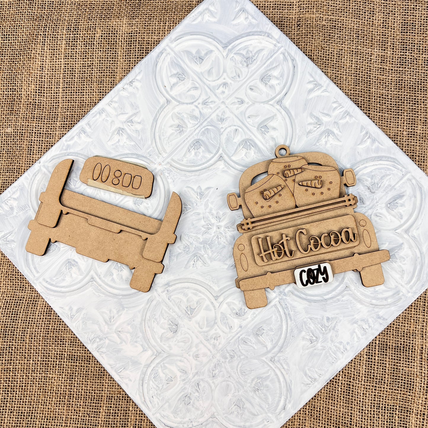 Create a delightful DIY kit with two wooden cutouts from Janet's Craft Corner's "Antique Truck Ornament" collection. One truck features "Hot Cocoa" accompanied by mugs on the bed, and the other has a blank sign, ideal for personalizing your Christmas décor. Both pieces maintain their natural wood finish, highlighting their vintage charm against a textured background.