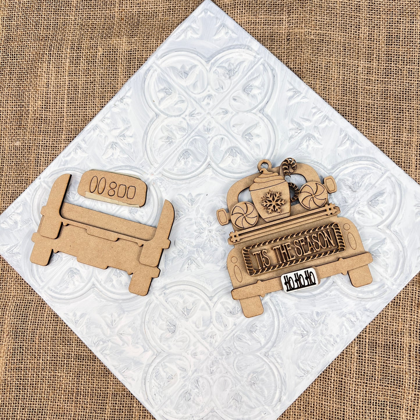 On a textured white surface, two wooden craft ornaments from Janet's Craft Corner are showcased as ideal additions to your Christmas décor. One ornament mimics the design of an antique truck with "000 800" elegantly displayed on the tailgate, while the other boasts festive messages such as "Tis the Season" and "Ho Ho Ho." The scene is set against a backdrop of burlap fabric.